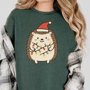Op de afbeelding: Donkergroen T-shirt met een cartoonegel met een kerstmuts en kerstverlichting. De egel heeft een cr&egrave;mekleurig lichaam, bruine stekels en een lachend gezicht. Het T-shirt heeft een relaxte pasvorm.