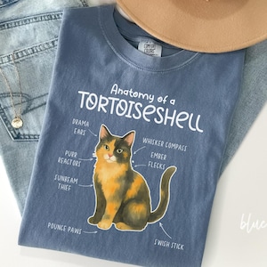 Pode incluir: Uma camiseta azul jeans com o texto "Anatomy of a Tortoiseshell" e um gráfico de um gato tartaruga. A camisa também apresenta um chapéu bege e um par de jeans.