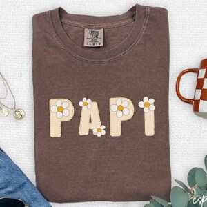May include: A brown Comfort Colors t-shirt with the word "PAPI" in cream-colored letters with daisy accents. A checkered mug and a necklace are visible. The shirt is folded on a white surface.