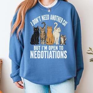 May include: A blue crewneck sweatshirt featuring a graphic of five cats of various colors and patterns. The white text above the cats reads I DON'T NEED ANOTHER CAT and the text below reads BUT I'M OPEN TO NEGOTIATIONS in a bold, sans-serif font.