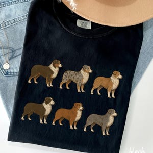 May include: Black t-shirt featuring embroidered images of six Australian Shepherds in various colors. The shirt has a crew neck and short sleeves. The tag reads "Comfort Colors". The word "black" is written in the bottom right corner.