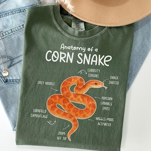 Puede incluir: Una camiseta verde oliva con el texto "Anatomy of a Corn Snake" y una ilustración detallada de una serpiente de maíz. La serpiente es naranja con manchas marrones y etiquetada con términos anatómicos humorísticos.