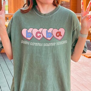 Peut inclure: Un t-shirt vert olive avec les lettres "bcba" dans des cœurs roses et du texte en dessous. La personne porte un bob vert assorti et des leggings noirs. L'arrière-plan comprend des tables de pique-nique et des bâtiments en bois.