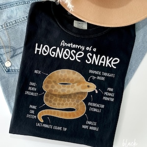 Pode incluir: Camiseta preta com um gráfico de uma cobra-nariz-de-porco marrom e bege e o texto "Anatomy of a Hognose Snake". A camisa também apresenta o texto "Comfort Colors" e "black".