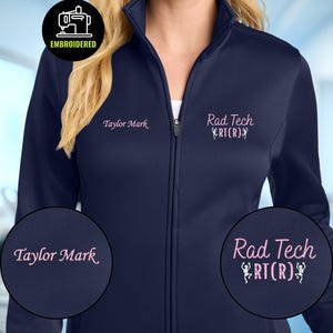 May include: A navy blue zip-up jacket featuring custom embroidery. The right chest displays the name Taylor Mark in pink script. The left chest features the text Rad Tech RT(R) in pink script with two small white skeleton graphics.