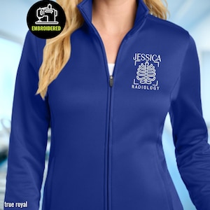 May include: A true royal blue full-zip jacket featuring a white embroidered design on the left chest. The design includes the name JESSICA above a stylized rib cage graphic and the word RADIOLOGY below. A circular black badge reads EMBROIDERED.
