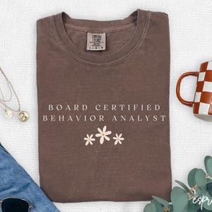 Puede incluir: Una camiseta marrón con el texto "BOARD CERTIFIED BEHAVIOR ANALYST" y tres pequeñas flores blancas. También se ven una taza a cuadros, gafas de sol y un collar.