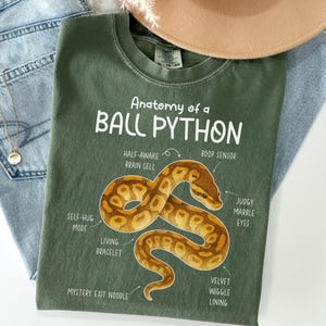 May include: An olive green t-shirt featuring the text "Anatomy of a Ball Python" and a diagram of a snake. The diagram labels parts of the snake with humorous terms like "Boop Sensor" and "Judgy Marble Eyes". The shirt is laid flat on a pair of blue jeans.