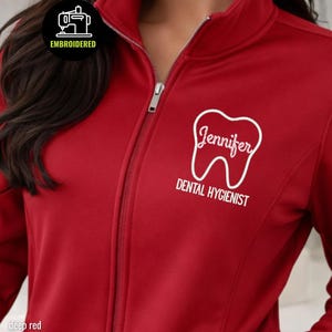 May include: Deep red zip-up jacket with white embroidered text and a tooth graphic. The text reads "Jennifer, Dental Hygienist." The jacket has a high collar and a silver zipper. The upper left corner has a small embroidered logo that says "EMBROIDERED".