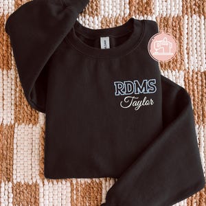 Peut inclure: Un sweat-shirt noir à manches longues et col rond avec du texte brodé sur la poitrine gauche. Le texte affiche 'RDMS' dans une police bloc bleu clair au-dessus du nom 'Taylor' en écriture cursive blanche. Un petit logo circulaire est visible.