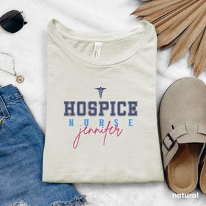 May include: A beige t-shirt with the text "HOSPICE NURSE jennifer" in blue and pink lettering. A medical symbol is above the text.