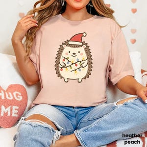 Op de afbeelding: Licht perzikkleurig T-shirt met een cartoonegel met een kerstmuts en omwikkeld met kerstverlichting. De tekst "heather peach" is zichtbaar. Het shirt wordt gecombineerd met een blauwe jeans.