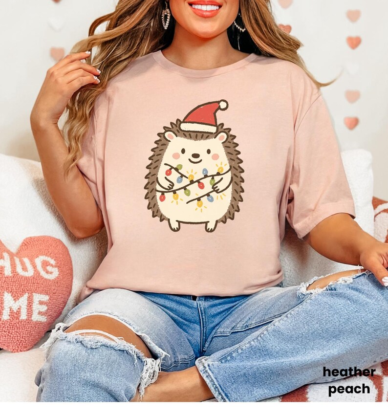 Op de afbeelding: Licht perzikkleurig T-shirt met een cartoonegel met een kerstmuts en omwikkeld met kerstverlichting. De tekst "heather peach" is zichtbaar. Het shirt wordt gecombineerd met een blauwe jeans.