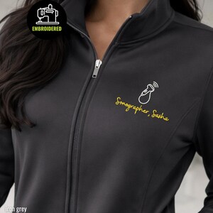 Puede incluir: Una chaqueta gris hierro con cremallera que presenta un icono de sonda de ultrasonido blanco bordado y el texto amarillo Sonographer, Sasha. Una insignia circular negra en la esquina superior izquierda muestra una máquina de coser y EMBROIDERED.