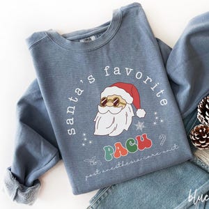 Könnte beinhalten: Ein blaues Sweatshirt mit dem Text "santa's favorite PACU" und einer Grafik von Santa Claus mit Sonnenbrille. Der Weihnachtsmann befindet sich in der Mitte des Sweatshirts. Das Sweatshirt ist auf einer blauen Jeans gefaltet.