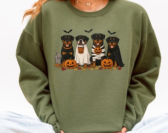 May include: An olive green crewneck sweatshirt featuring a graphic print of four Rottweiler dogs dressed in Halloween costumes, including a ghost, a mummy, and a vampire, alongside jack-o'-lantern pumpkins, fallen autumn leaves, and bats.