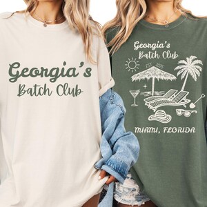 May include: Two t-shirts, one cream and one olive green, with the text "Georgia's Batch Club". The green shirt has a beach scene graphic with the text "Miami, Florida". Both shirts have rolled sleeves.