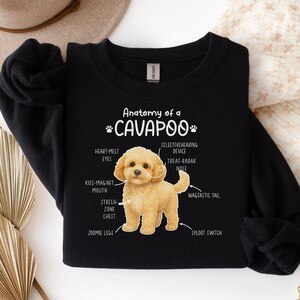 Op de afbeelding: Zwarte crewneck sweatshirt met de tekst "Anatomy of a Cavapoo" en een diagram van een Cavapoo hond. Het diagram labelt de kenmerken van de hond, waaronder "Heart-Melt Eyes" en "Wagtastic Tail".