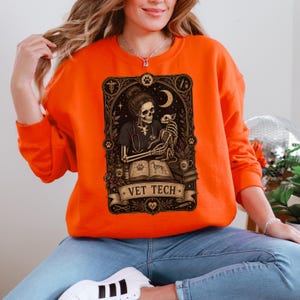 Puede incluir: Una sudadera de cuello redondo color naranja brillante con un estampado gráfico negro detallado de un esqueleto con estetoscopio que sostiene a un perro esqueleto. El diseño incluye un libro abierto, símbolos médicos y el texto VET TECH.