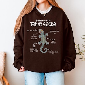 May include: A dark brown sweatshirt featuring the text "Anatomy of a Tokay Gecko" with an illustrated gecko and labeled body parts. The gecko is light blue with orange polka dots. The sweatshirt has long sleeves.