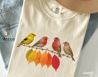 May include: Ivory-colored t-shirt featuring a colorful bird illustration. The design shows four birds perched on a branch with autumn leaves. The shirt is made of a soft material and has a tag that says "Comfort Colors".