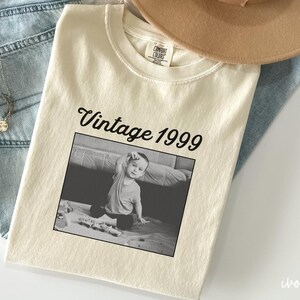 Può includere: T-shirt color crema con la scritta "Vintage 1999" sopra una foto in bianco e nero di un bambino piccolo. La maglietta è appoggiata su jeans blu e un cappello beige. È visibile anche una collana d'argento con un ciondolo.