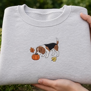 Könnte beinhalten: Ein hellgraues Sweatshirt mit Rundhalsausschnitt und einer zentralen Stickerei, die einen Beagle-Hund neben einem orangefarbenen Kürbis zeigt, ergänzt durch zwei kleine Herbstblätter in Gelb- und Brauntönen, mittig auf der Brust.