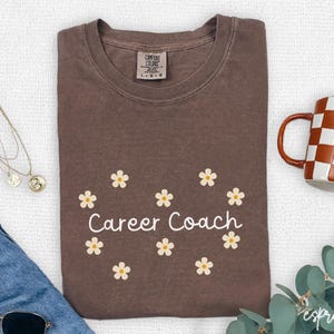 May include: A brown t-shirt with the text "Career Coach" surrounded by small daisy graphics. A checkered mug, sunglasses, and a necklace are also visible.