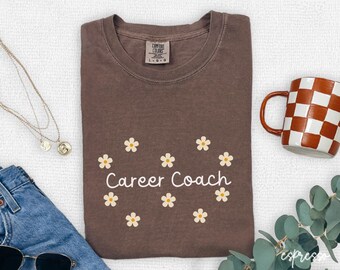 May include: A brown t-shirt with the text "Career Coach" and daisy graphics. A checkered mug, sunglasses, and a necklace are also visible.
