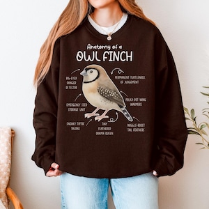 Puede incluir: Una sudadera marrón oscuro con el texto "Anatomy of a Owl Finch" y una ilustración de un pájaro. El pájaro es beige, marrón y blanco, con etiquetas como "polka-dot wing warmers" y "tiny feathered drama queen".