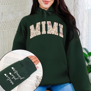 以下が含まれることがあります： 胸に花柄のデザインで「MIMI」の文字が入った濃い緑色のパーカー。左袖には「Amelia」、「John」、「Jennifer」の名前がハートのシンボルとともに縦にプリントされています。
