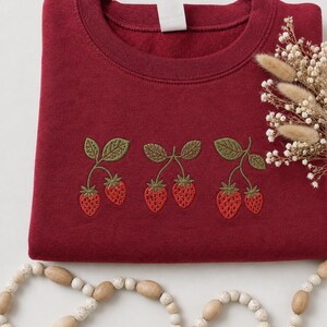 May include: A deep red crewneck sweatshirt featuring an embroidered design of three pairs of strawberries with green leaves and stems. The embroidery is centered on the chest, showcasing detailed red fruit textures and olive green foliage.
