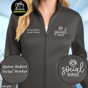 May include: An iron grey full-zip jacket featuring white embroidery. The right chest displays the text Quinn Robert Social Worker. The left chest features a graphic of hands holding a group of people with the text social WORKER below it.