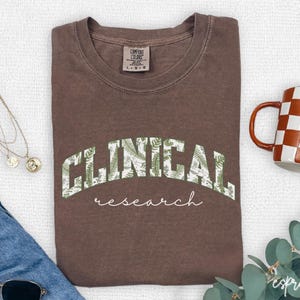 May include: A brown t-shirt with a green and white graphic that says "CLINICAL research" in a vintage collegiate style.