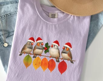 May include: A lilac Comfort Colors t-shirt featuring a graphic of four birds wearing Santa hats perched on a branch with colorful leaves. The shirt is folded on a denim surface, with a hat and necklace visible.