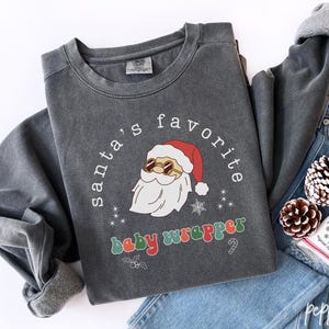 Puede incluir: Sudadera gris oscuro con el texto "santa's favorite baby wrapper" y un gráfico de Papá Noel con gafas de sol. La sudadera está doblada sobre unos vaqueros azules, con piñas y una pequeña bolsa.