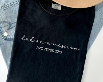 May include: Black t-shirt with white script reading "dad on a mission" and "Proverbs 22:6". The shirt is folded, with a tan hat and blue jeans visible. The t-shirt is on a white surface.