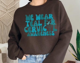 Może przedstawiać: Brązowa bluza z nadrukiem w kolorze turkusowym, na którym widnieje napis "We Wear Teal For Cervical Awareness". Nadruk zawiera turkusową wstążkę i symbol macicy.