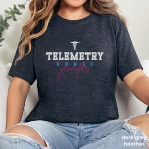 Könnte beinhalten: Ein dunkelgraues Heather-T-Shirt mit dem Text "Telemetry Nurse Jennifer" in weißer und blauer Schrift. Ein weißes Caduceus-Symbol befindet sich über dem Text.