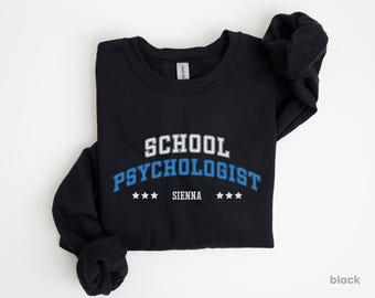 Puede incluir: Una sudadera negra con las palabras "SCHOOL PSYCHOLOGIST" en letras blancas y azules. Debajo del texto hay tres estrellas blancas y la palabra "SIENNA". La sudadera es de cuello redondo.
