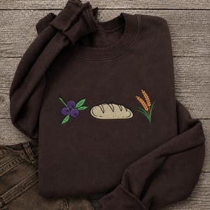 May include: A dark brown crewneck sweatshirt featuring an embroidered design on the chest. The embroidery depicts a cluster of three purple blueberries with green leaves, a single beige loaf of bread, and a stalk of golden wheat with green leaves.
