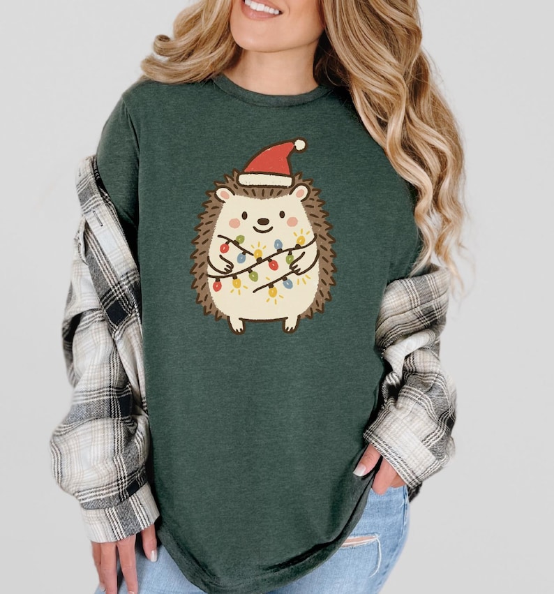 Op de afbeelding: Donkergroen T-shirt met een cartoonegel met een kerstmuts en kerstverlichting. De egel heeft een cr&egrave;mekleurig lichaam, bruine stekels en een lachend gezicht. Het T-shirt heeft een relaxte pasvorm.