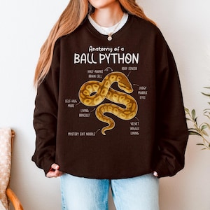 Könnte beinhalten: Ein dunkelbraunes Sweatshirt mit dem Text "Anatomy of a Ball Python" und einer Abbildung einer Schlange. Die Abbildung enthält Beschriftungen wie "Self-Hug Mode" und "Judgy Marble Eyes."