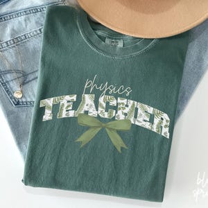 May include: A green t-shirt with the text "Physics Teacher" in white letters with a leafy pattern. A green bow is below the text.