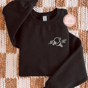 May include: Black crewneck sweatshirt with white embroidered ping pong paddles and the initials "MK". The sweatshirt is made of a soft material and is folded on a patterned surface. The word "black" is visible at the bottom.