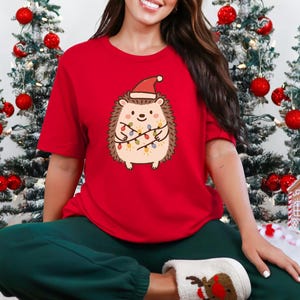 Op de afbeelding: Rood T-shirt met korte mouwen met een cartoonegel met een kerstmuts en omwikkeld met kerstverlichting. De egel heeft een lachend gezicht. De achtergrond bevat kerstbomen met rode ballen.