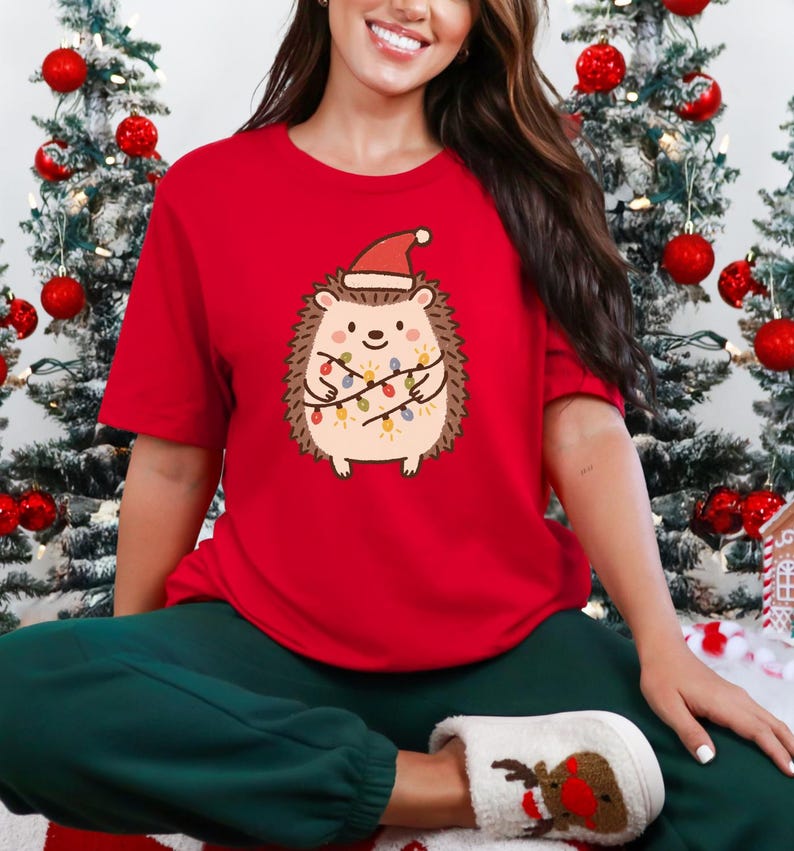 Op de afbeelding: Rood T-shirt met korte mouwen met een cartoonegel met een kerstmuts en omwikkeld met kerstverlichting. De egel heeft een lachend gezicht. De achtergrond bevat kerstbomen met rode ballen.