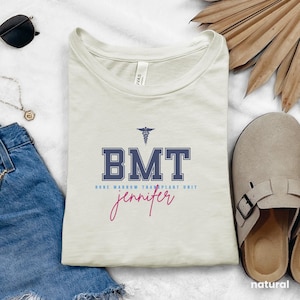 May include: A beige t-shirt with a blue and white graphic that says "BMT Bone Marrow Transplant Unit" with a medical symbol above the text. The name "jennifer" is written in pink cursive below the graphic.