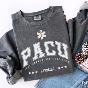 May include: A charcoal grey sweatshirt with the text "PACU POST ANESTHESIA CARE UNIT CAROLINE" in white. The sweatshirt has a medical symbol and three stars. The sleeves are rolled up, and it's paired with blue jeans and pine cones.