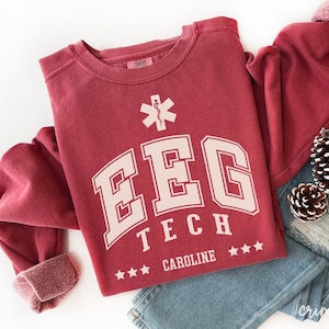 May include: A maroon-colored sweatshirt with the text "EEG TECH CAROLINE" in white block letters. The design includes a medical symbol and three stars. The sweatshirt is folded on top of blue jeans.